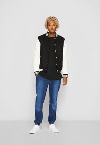 Varsity jacket with a black body and white sleeves, featuring striped ribbed cuffs. Paired with blue jeans and white sneakers.