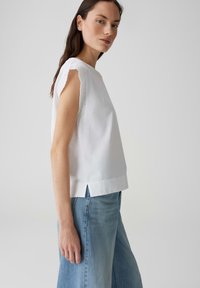 Witte katoenen top met geborduurde kantaccenten op de mouwen, rechte snit en zijsplitten. Gemaakt met lichtblauwe wijde spijkerbroeken.