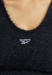 Svart, fuzzy Reebok crop-top med V-hals design. Har brodert logo i hvitt på venstre brystområde. Myk tekstur.