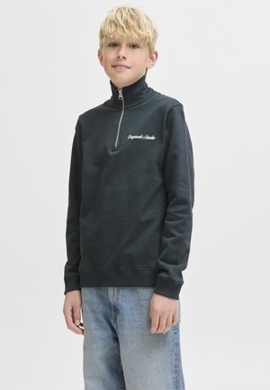 Garçon blond portant un sweat-shirt noir à fermeture éclair quart de zip avec le texte "Originals Studio" et un jean bleu clair, debout devant un fond blanc uni.