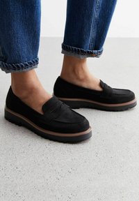 Black suede loafers with a stitched toe design, a raised seam across the top, and a thick rubber sole. Worn with rolled cuff denim.