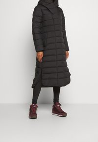 Giacca imbottita nera con cappuccio, design fino al ginocchio e tasche laterali. Indossata con leggings neri e calzature color borgogna.