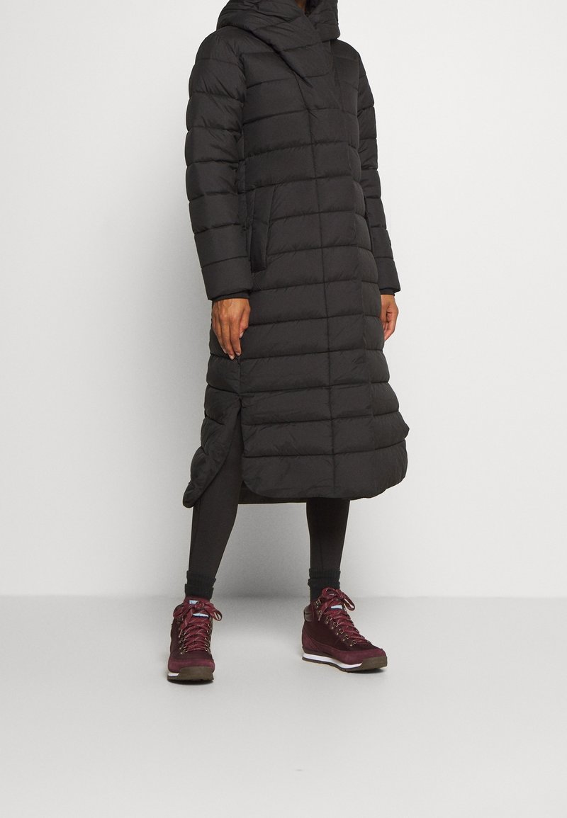 Giacca imbottita nera con cappuccio, design fino al ginocchio e tasche laterali. Indossata con leggings neri e calzature color borgogna.