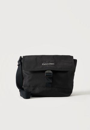 Czarna torebka na ramię Calvin Klein z przednią klapą, zapięciem na klamrę i regulowanym paskiem na białym tle.