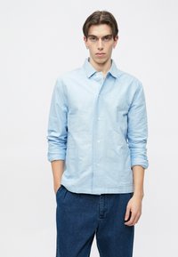 Hele sinine nupuga särk, mis on valmistatud pehmest kangast, ilma kraeta, pika varrukaga ja kahe esiosa taskuga.