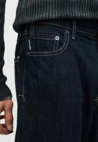 Κοντινό πλάνο ενός ατόμου που φοράει σκούρο μπλε τζιν με αντιθετική ραφή και μια μικρή μπροστινή τσέπη με την ετικέτα "JACK&JONES".