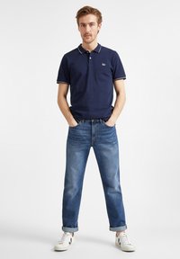 Granatowa koszulka polo z białymi wykończeniami, dżinsy z podwiniętymi nogawkami oraz białe sneakersy z czarnymi akcentami. Standardowy krój i casualowy design.