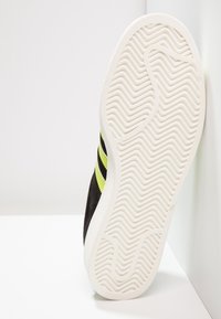 Svart mockasneaker med vit gummisula, med tre limegröna ränder och ett texturerat zigzag-mönster på sulan.