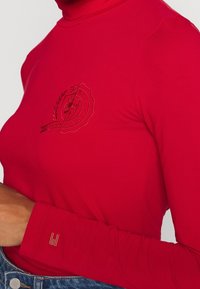 Red long-sleeve top with a high neck, featuring an embossed logo on the chest and a small gold accent on the sleeve. Smooth, fitted texture.