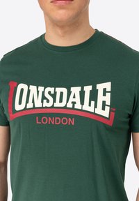 Žalios spalvos medvilninė marškinė su ryškiu baltu ir raudonu "LONSDALE" logotipu, po kuriuo yra "LONDON", apvaliu kaklu ir trumpomis rankovėmis.