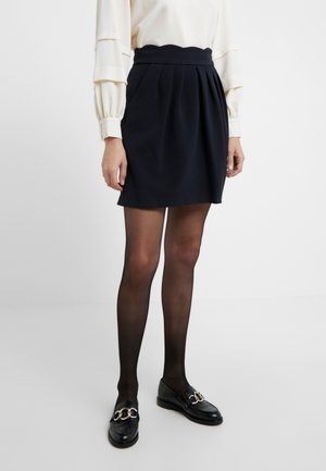 Kvinde iført en sort plisseret mininederdel, gennemsigtige sorte strømpebukser, sorte loafers med sølvkædedetaljer og en cremefarvet langærmet bluse.