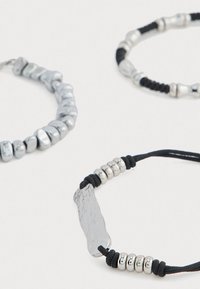 Tres pulseras de cuentas con cuentas en tonos plateados, un colgante de plata texturizado y acentos de cordón negro. Presentan tamaños y formas de cuentas variadas.
