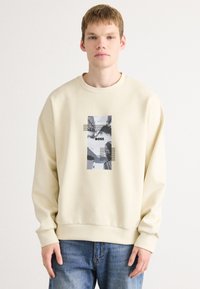 Kremfarget hoodie laget av myk stoff, med et grafisk trykk i svart og grått plassert på forsiden, med ribbestrikkede mansjetter og nederkant.