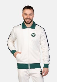 Weißer Zip-Jacke mit grünem Kragen und Bündchen, marineblauen Seitenstreifen und einem runden grünen Logo auf der Brust. Glatte Stoffoberfläche.