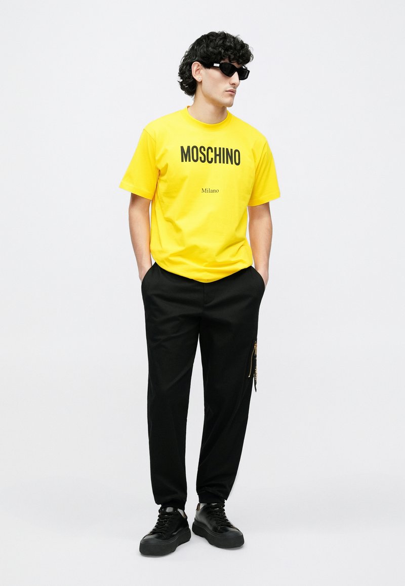 Gul bomuld T-shirt med "MOSCHINO Milano" i sort tryk, kombineret med sorte bukser med lynlås i siden og sorte sneakers.