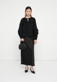 Crna bluzica s volanima i dekolteom, u kombinaciji s elegantnom crnom maksi suknjom. Model drži malu crnu torbicu s teksturom i nosi crne štikle.