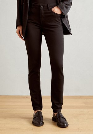 Persoon die zwarte strakke broek, zwarte instapschoenen en een bijpassend zwart jasje draagt, met één hand in de zak, staand op een houten vloer.