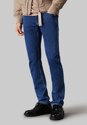 Jean en denim bleu avec une coupe droite, matériau premium, texture subtile et détails de couture blanche. Porté avec des chaussures noires et une veste beige.