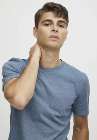 Ung mann i en blå t-skjorte som tar på baksiden av nakken, ser litt oppover mot en enkel lys bakgrunn.