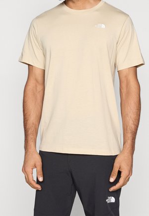 Mand iført en beige The North Face t-shirt med korte ærmer og sorte bukser med The North Face-logo på brystet og låret.
