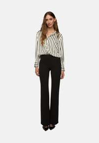 Donna in piedi con una camicia bianca a righe verticali nere, pantaloni neri a vita alta e scarpe nere a punta.