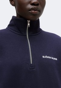 Gros plan d'une personne portant un haut zippé bleu marine avec un col montant et un logo blanc "BJÖRN BORG" sur la poitrine.