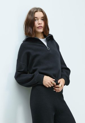 Black zip-up sweater made from soft material, featuring a high collar, long sleeves, and ribbed cuffs. Paired with matching dark pants.