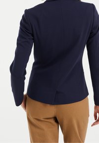 Tummansininen räätälöity bleiseri, jossa on istuva malli, rakenteelliset hartiat ja sileä pinta, yhdistetty vaaleanruskeisiin housuihin.