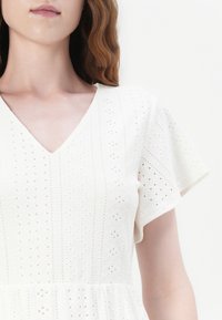 Femme à la peau claire et aux cheveux ondulés châtain clair portant une robe blanche en tissu ajouré avec de courtes manches volantées et un décolleté en V.
