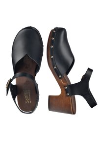 Zoccoli in pelle nera con punta aperta, tacco in legno e borchie in ottone lungo i lati. Presentano un design con ritagli e una cintura regolabile.