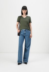 Maglietta verde oliva a V, jeans a vita alta blu in denim larghi, tacchi a punta neri, borsa a mano, sfondo bianco semplice.