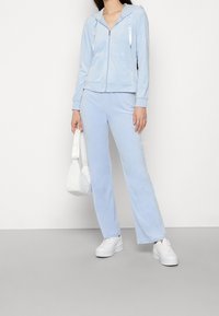 Ljusblå velourtracksuit med huva, dragkedja och matchande utsvängda byxor. Matchas med vita platåskor och en liten vit väska.