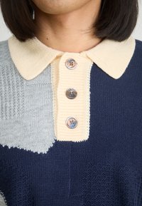 Πλεχτό πουλόβερ με γιακά σε ναυτικό μπλε, ανοιχτό γκρι και κρεμ τμήματα. Διαθέτει τρία στρογγυλά κουμπιά και ποικιλία υφής στα σχέδια.