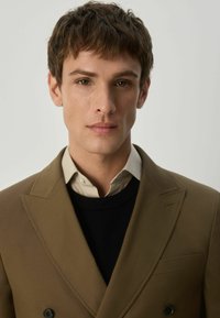 Brūna dubultā krūšu žakete, izgatavota no gludas auduma, virs melnas džemperis un gaišas krekla. Īpašības: nogrieztie klāji un priekšējās kabatas.