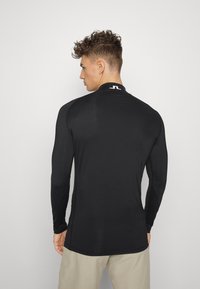 Crna košulja s dugim rukavima izrađena od glatkog materijala. Ima visoki ovratnik i logo na leđima. Slim fit dizajn bez uzoraka.