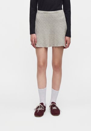 Personne portant une mini-jupe beige en maille torsadée, un haut noir à manches longues, des chaussettes blanches et des chaussures bordeaux à lacets, debout devant un fond blanc.