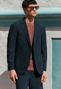 Marinefarvet tekstureret blazer med notch-lapper, enkelt knaplukning og to lommer, lagdelt over en brun T-shirt. Model iført solbriller.