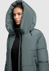 Giacca puffa in verde opaco con un grande cappuccio strutturato. Presenta tasche con zip e una zipper frontale, realizzata in un materiale liscio e leggero.