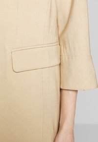 Beige jacket with a smooth texture, featuring a folded sleeve and a rectangular patch pocket on the left side.