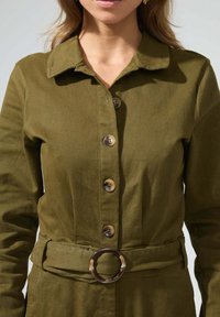 Robe chemise en coton vert avec col, boutons sur le devant et une ceinture avec une boucle circulaire à motif. Texture lisse et coupe ajustée.