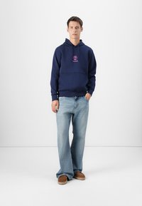 Navy sweatshirt with a hood and front kangaroo pocket, featuring a pink Timberland logo. Worn with light blue wide-leg jeans and brown shoes.