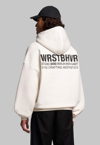 Felpa bianca oversize con una grande stampa grafica nera sul retro. Presenta un cappuccio con cordino e polsini a coste. Indossata con pantaloni neri.