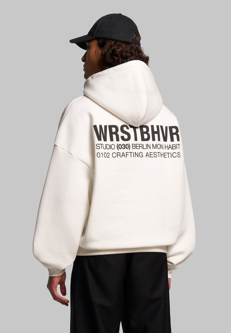Felpa bianca oversize con una grande stampa grafica nera sul retro. Presenta un cappuccio con cordino e polsini a coste. Indossata con pantaloni neri.