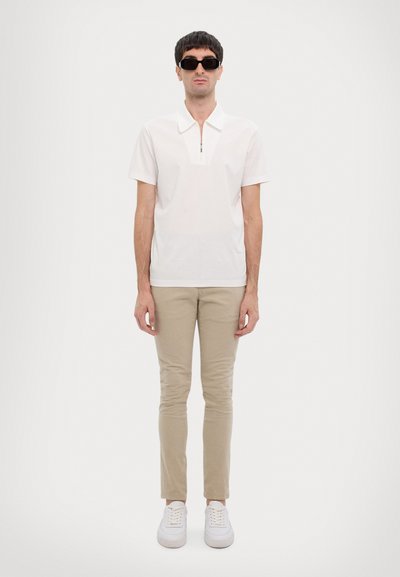 Polo blanc à manches courtes avec col zippé, pantalon slim beige et baskets blanches. Le modèle porte des lunettes de soleil noires et se tient devant un fond clair.