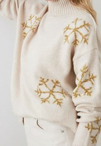 Cremfarbener Rollkragenpullover mit goldenen Glitzer-Schneeflockenmustern, getragen von einer Person mit langen blonden Haaren, kombiniert mit weißen Hosen.