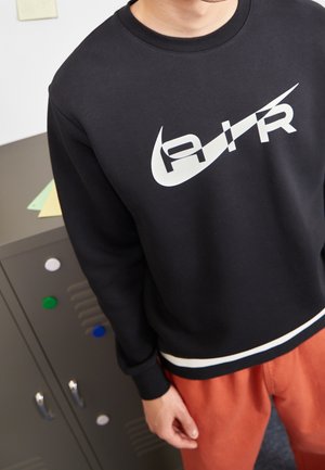 Osoba ubrana w czarną bluzę z kapturem z białym stylizowanym logo "AIR" oraz pomarańczowe spodnie, stojąca obok szarej szafki.