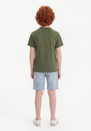 Kind mit lockigen roten Haaren steht mit dem Rücken zum Betrachter, trägt ein olivgrünes T-Shirt, helle Jeansshorts und weiße Slip-On-Schuhe auf weißem Hintergrund.