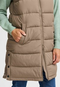 Gilet en duvet beige avec un design matelassé horizontal, fermeture éclair à l'avant et poches latérales. Dispose de fermetures à pression à l'ourlet et d'une texture douce.