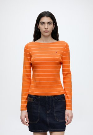 Mujer con cabello largo y oscuro que lleva una camiseta de manga larga naranja con rayas y una falda vaquera oscura, de pie frente a un fondo blanco liso.