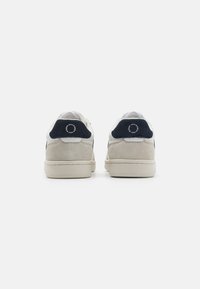Weiße und marineblaue Sneakers mit einem Wildleder-Rückenteil, strukturierten Außensohlen und kontrastierenden Nähten. Die Rückansicht hebt das Logo an der Ferse hervor.